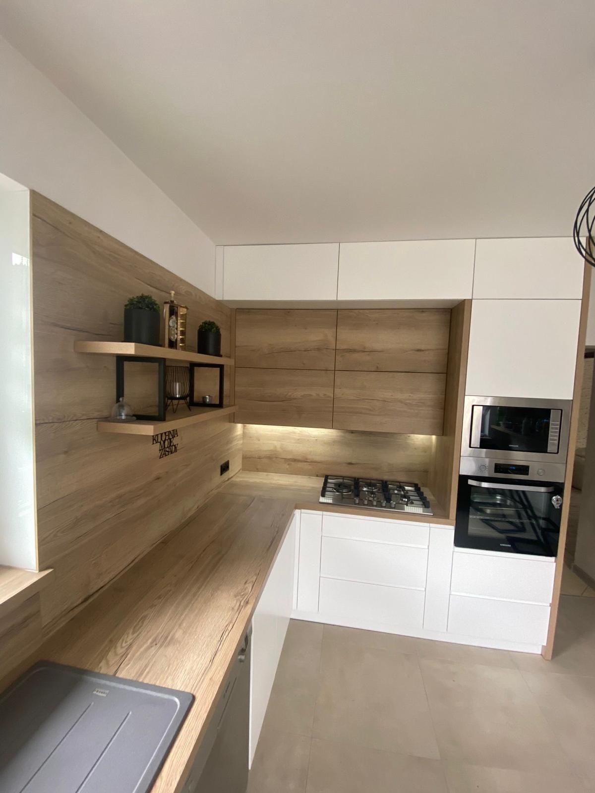 Moderne L-förmige Küche mit weißen Schränken, hellen Arbeitsplatten und Spritzschutz in Holzoptik sowie eingebauten Geräten.