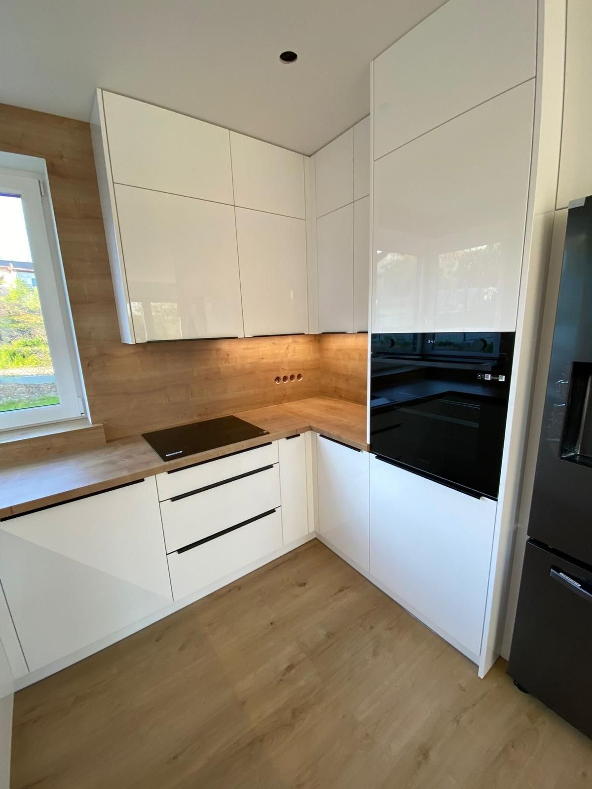 Eine moderne Küchenecke mit weißen Hochglanzschränken, hellen Arbeitsplatten in Holzoptik und einem eingebauten schwarzen Backofen.