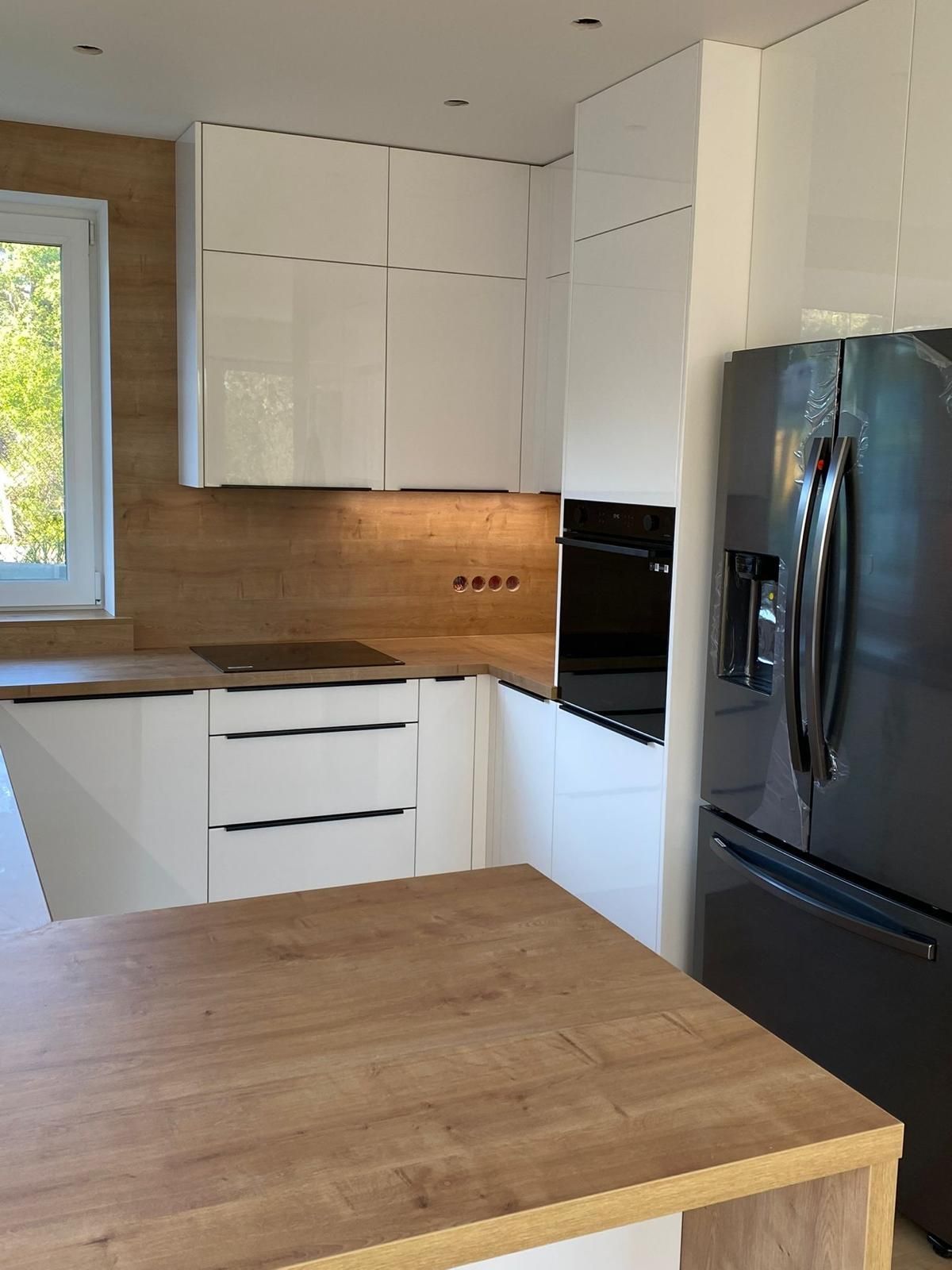 Moderne Küche mit weißen Hochglanzschränken, Arbeitsplatten und Spritzschutz in Holzoptik sowie einem schwarzen Kühlschrank.