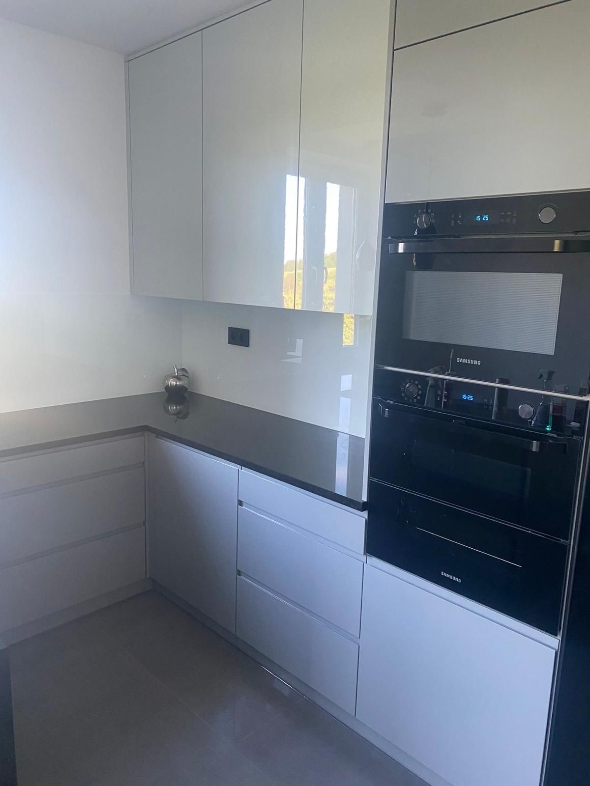 Eine moderne Küchenecke mit hellgrauen Hochglanzschränken, einer dunklen Arbeitsplatte und einem eingebauten schwarzen Backofen und einer Mikrowelle.