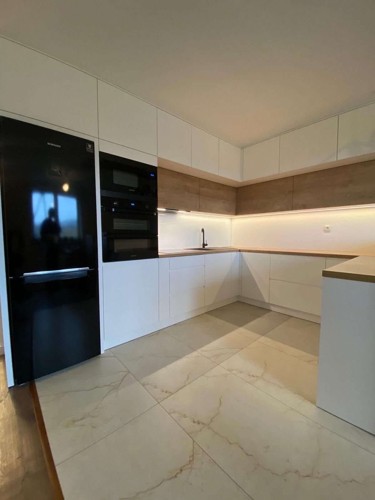 Moderne U-förmige Küche mit weißen Schränken, hellen Holzelementen, Unterbaubeleuchtung und einem großen schwarzen Kühlschrank.