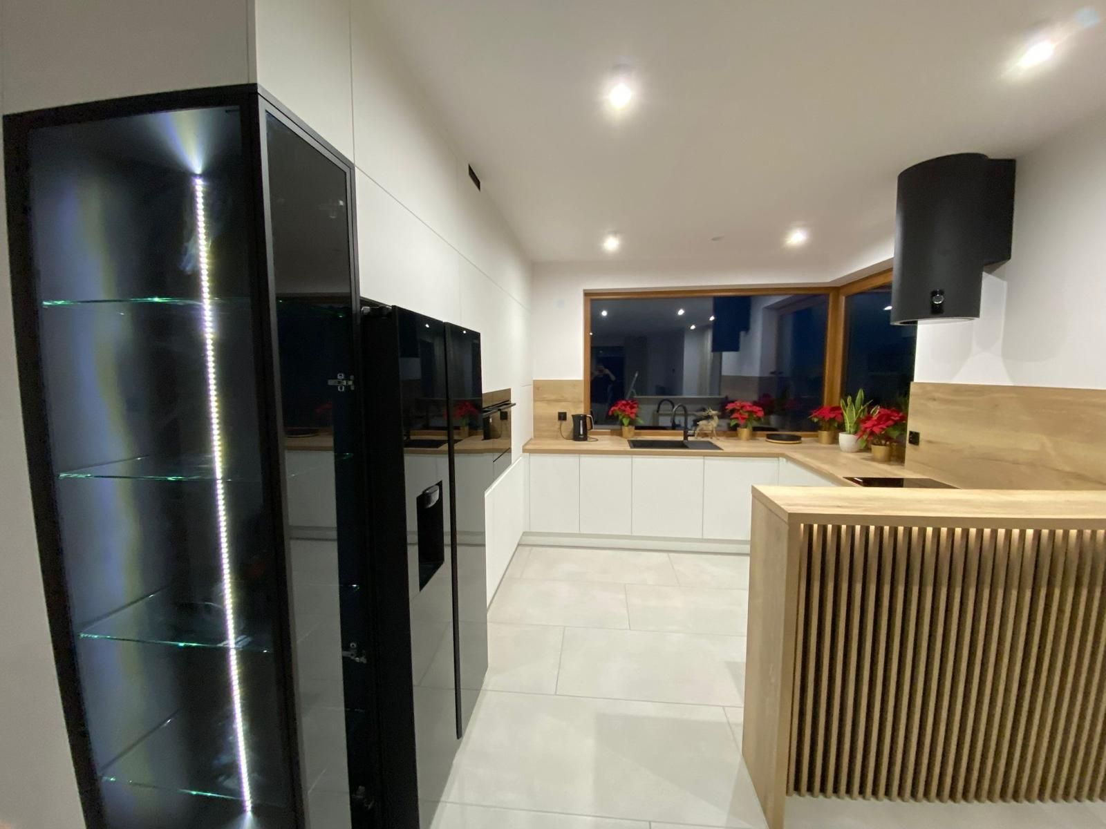 Moderne Küche mit weißen Schränken, einem schwarzen Kühlschrank, einer beleuchteten Glasvitrine und einer Kücheninsel aus Lattenholz.