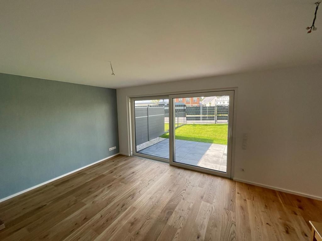 Leerer Raum mit Holzboden, einer salbeigrünen Akzentwand und einer Glasschiebetür, die auf eine Terrasse und in den Garten führt.