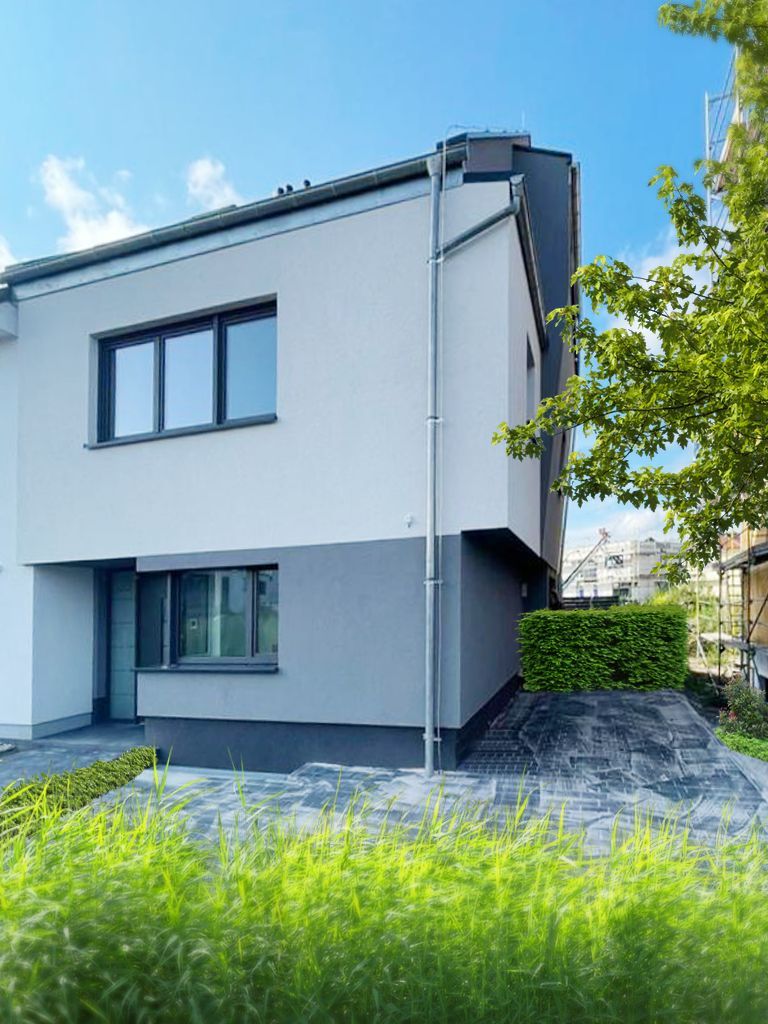 Ein modernes zweistöckiges Haus mit hellgrauen oberen und dunkelgrauen unteren Wänden, gelegen neben einem gepflasterten Bereich und einer grünen Hecke.
