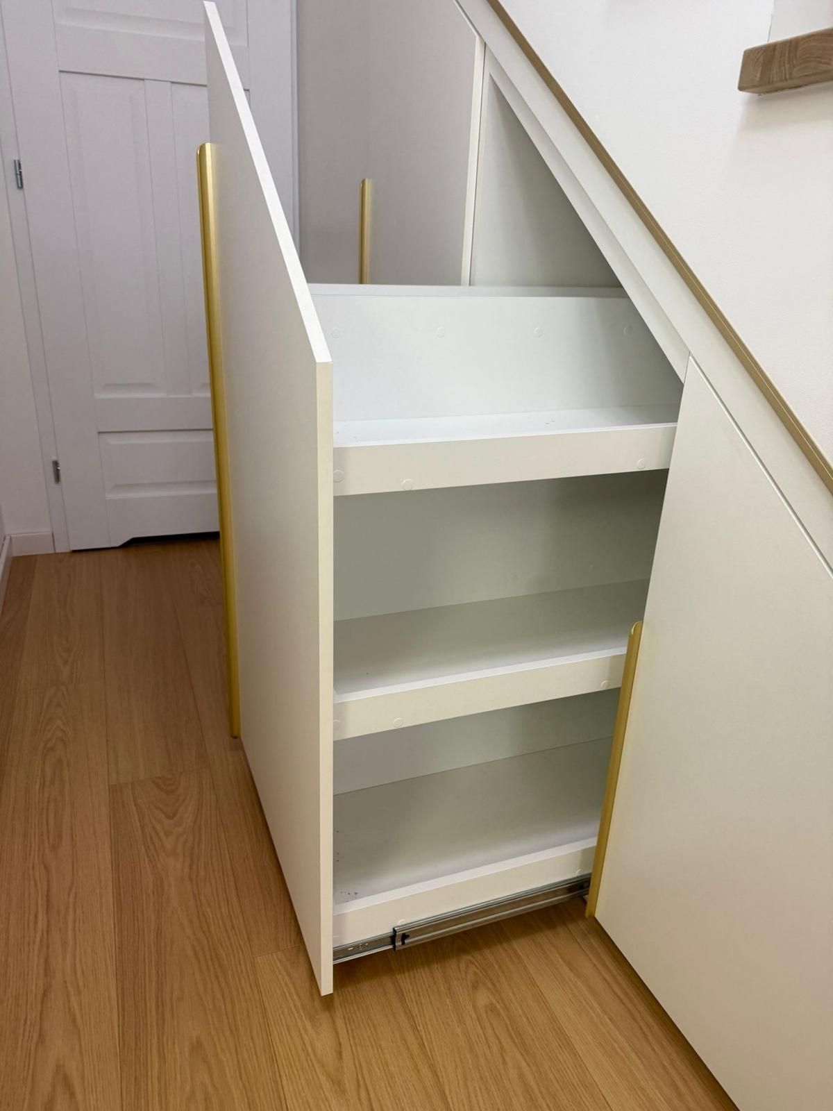 Ein weißer, ausziehbarer Untertreppenschrank mit drei Regalböden und goldener vertikaler Zierleiste, der auf einem Boden in Holzoptik steht.