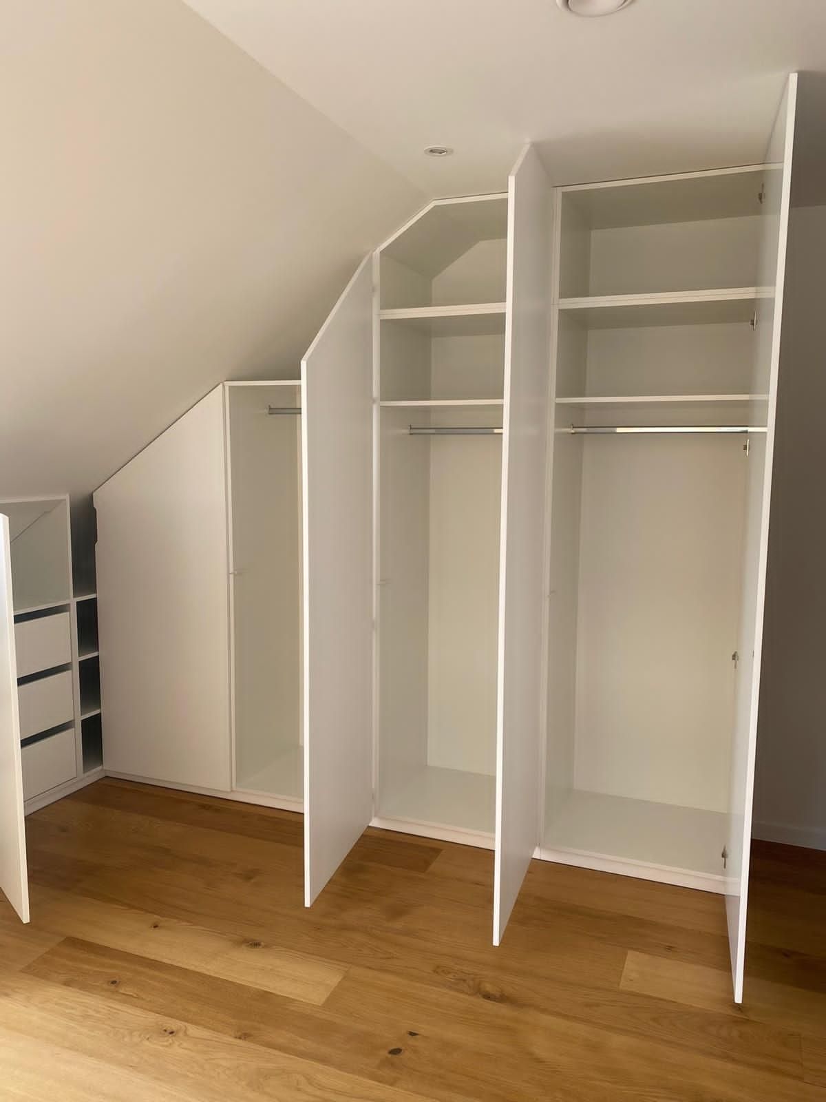 In einem Zimmer mit schräger Decke befinden sich mehrere weiße, maßgefertigte Einbauschränke mit offenen Regalen und Kleiderstangen.