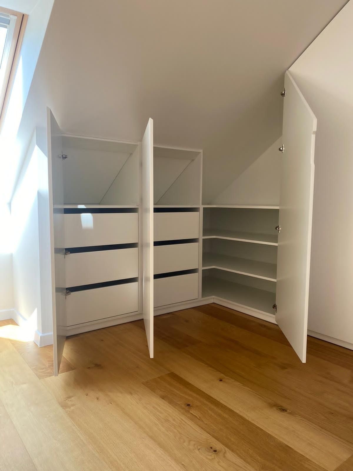 Eingebaute weiße Aufbewahrungsschränke mit Schubladen und offenen Regalen wurden unter einer schrägen Dachbodendecke auf einem Holzboden installiert.