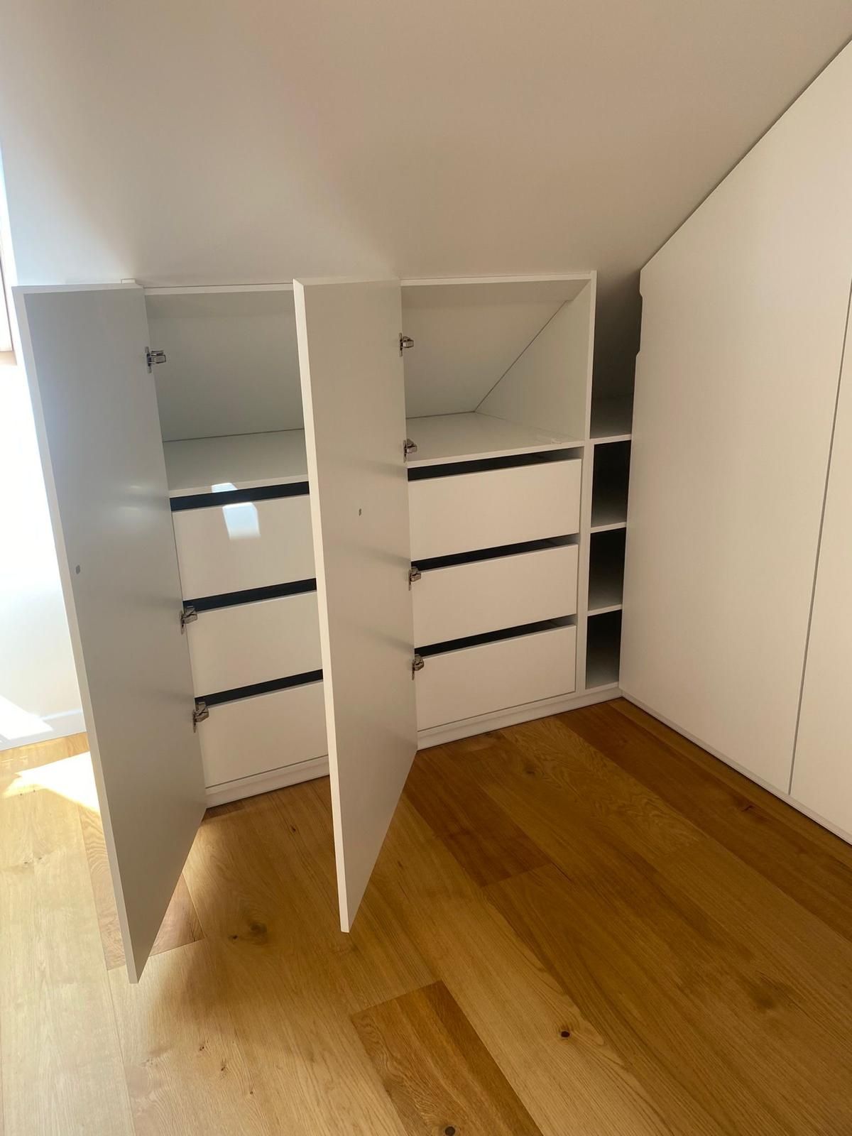 Maßgefertigte, weiße Einbauschränke mit offenen Türen und Schubladen, die in einen schrägen Dachboden mit hellem Holzboden integriert sind.