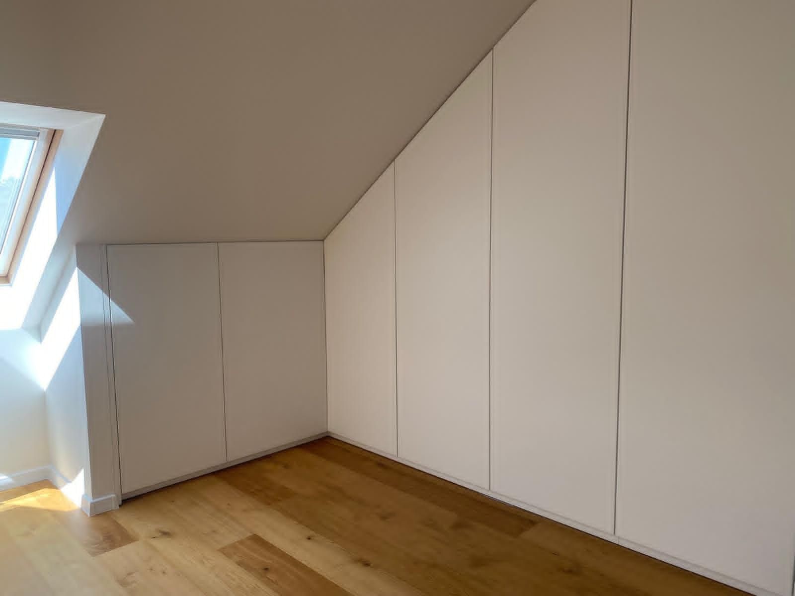 In einem Raum mit hellem Holzboden fügen sich weiße Einbauschränke mit schrägen Türen harmonisch in die Dachschräge ein.