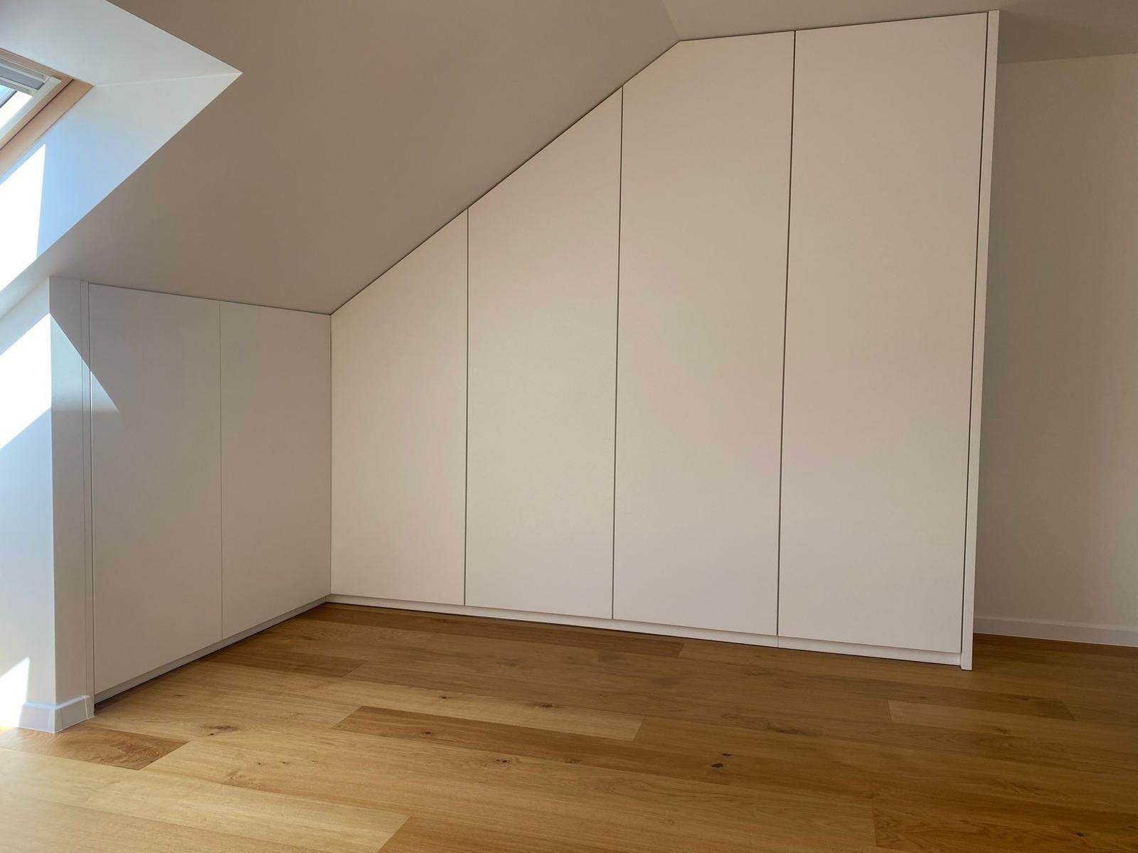 Weiße, maßgefertigte Kleiderschränke wurden entlang einer schrägen Dachbodenwand über hellem Holzboden installiert.