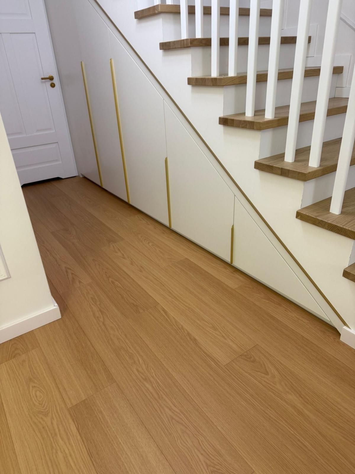 Weiße Unterschränke mit goldenen, vertikalen Griffen neben einer Holztreppe und hellem Holzboden.