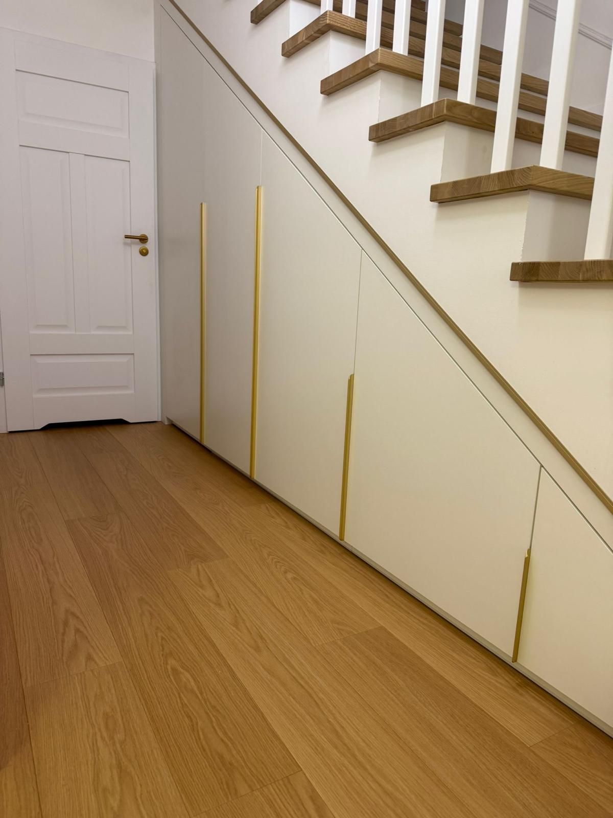 Eingebaute weiße Aufbewahrungsschränke mit goldenen vertikalen Griffen befinden sich unter einer Holztreppe in einem Flur.
