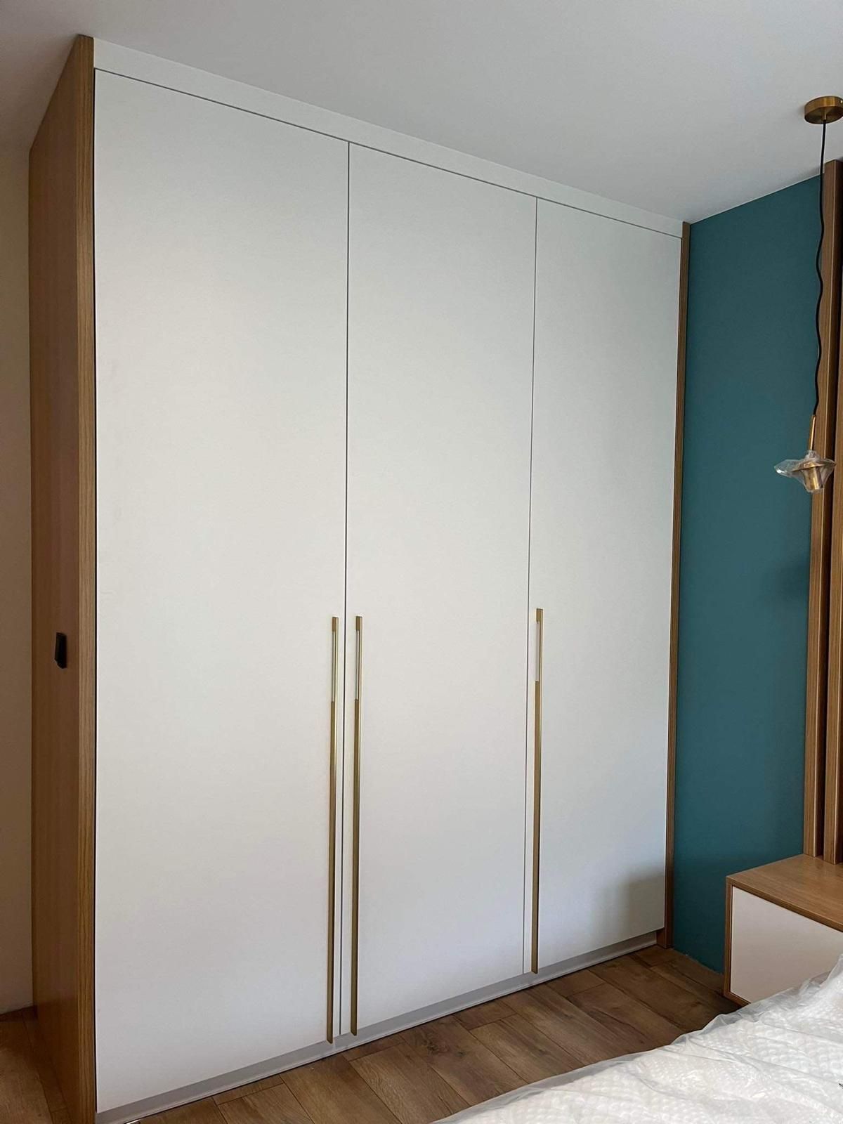 Ein hoher, moderner weißer Kleiderschrank mit langen, goldfarbenen vertikalen Griffen, der neben einer dunkelgrünen Akzentwand steht.