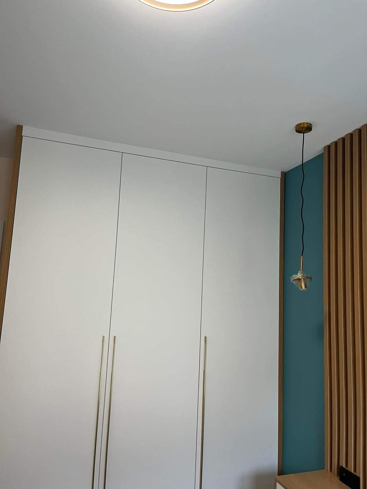 Hohe, weiße Kleiderschranktüren vor einer türkisfarbenen Wand mit vertikalen Holzlamellen und einer hängenden Messingpendelleuchte.