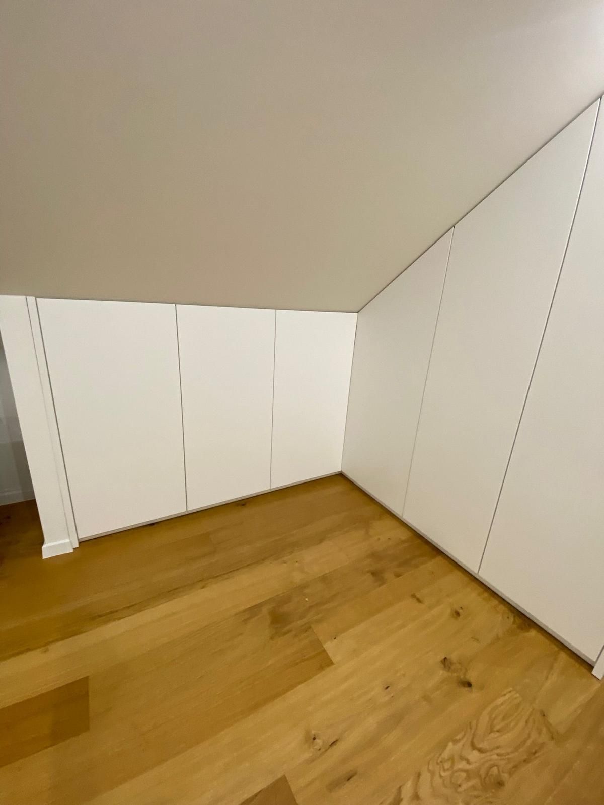 Eingebaute weiße Stauschränke unter einer schrägen Dachbodendecke mit hellem Holzboden.