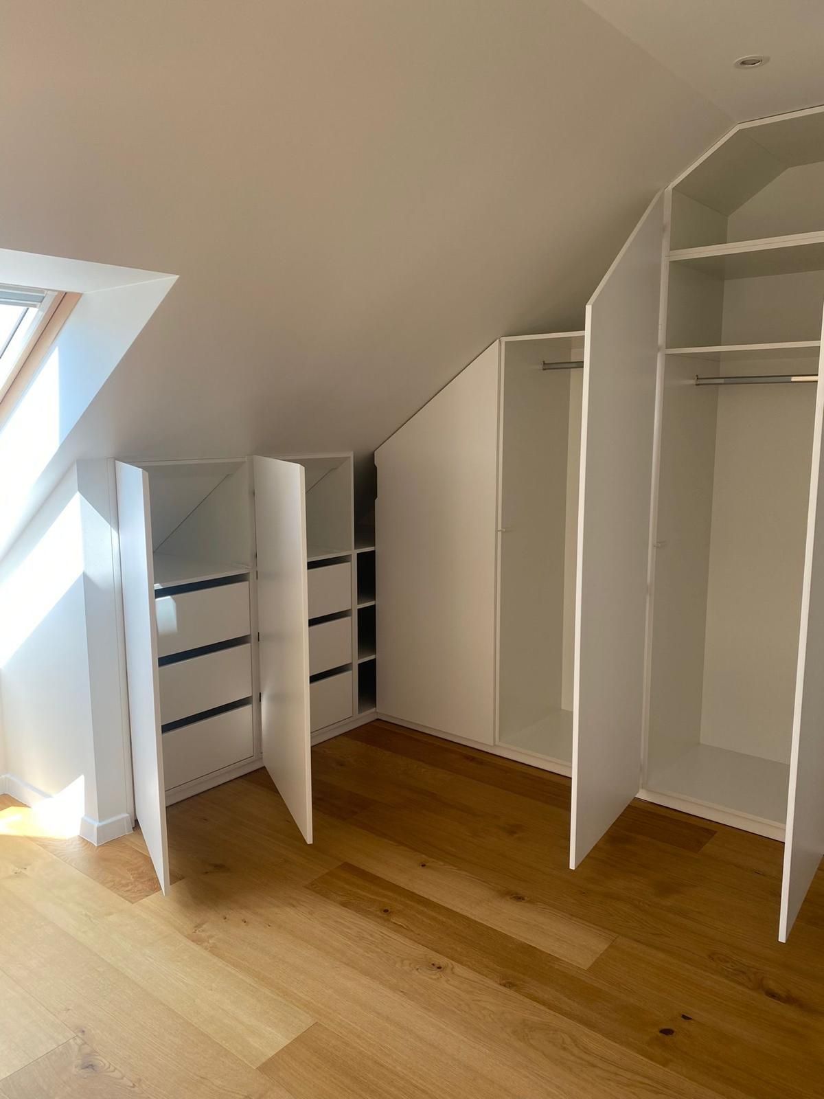 Eingebaute weiße Kleiderschränke in einem Dachzimmer mit Dachschrägen, offenen Türen, Regalen und hellem Holzboden.