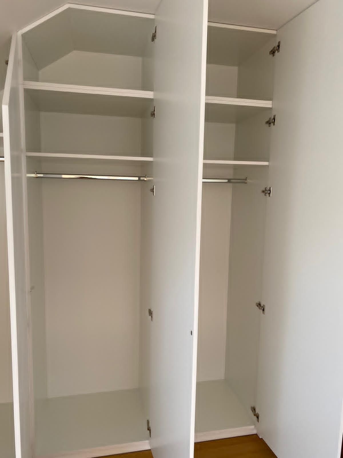 Ein weißer Kleiderschrank mit drei Abteilen, ausgestattet mit Regalböden, Kleiderstangen und einer schrägen Oberseite auf der linken Seite.