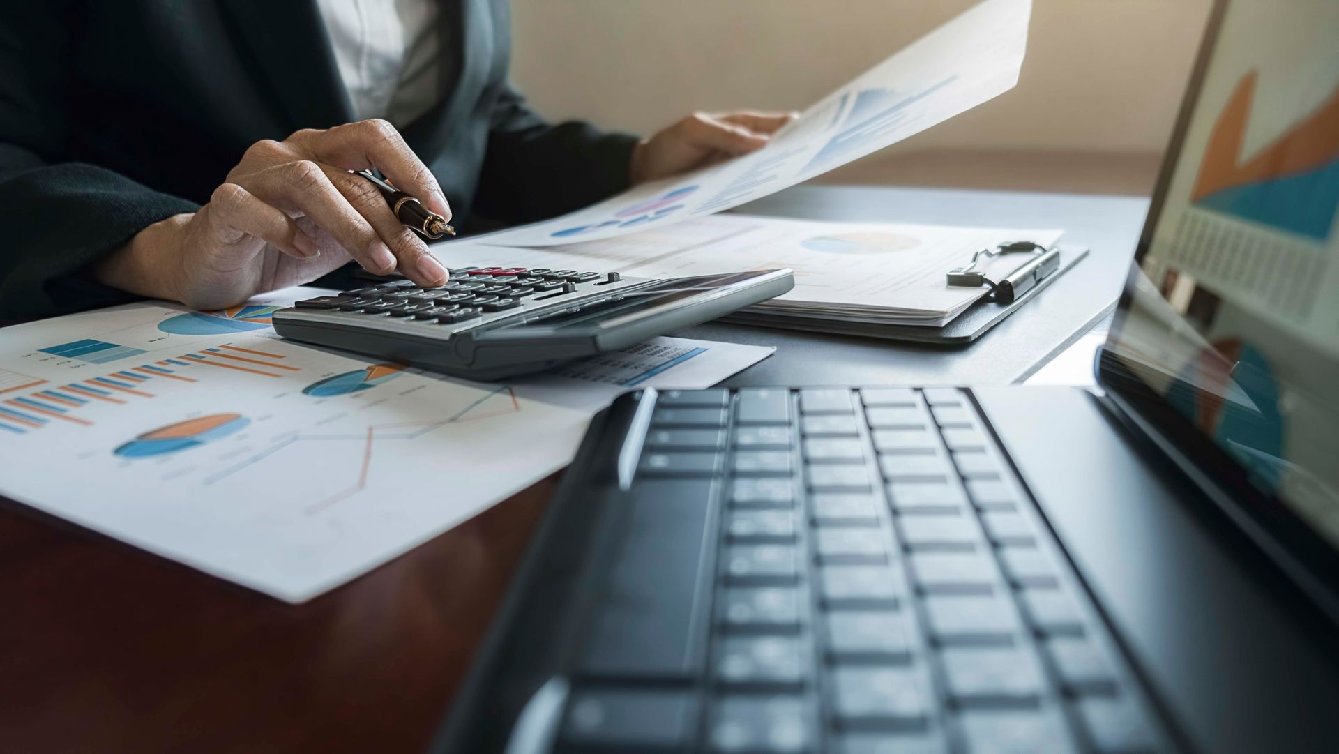 Person, die mit Taschenrechner, Dokumenten und Laptop auf einem Schreibtisch Finanzen berechnet.