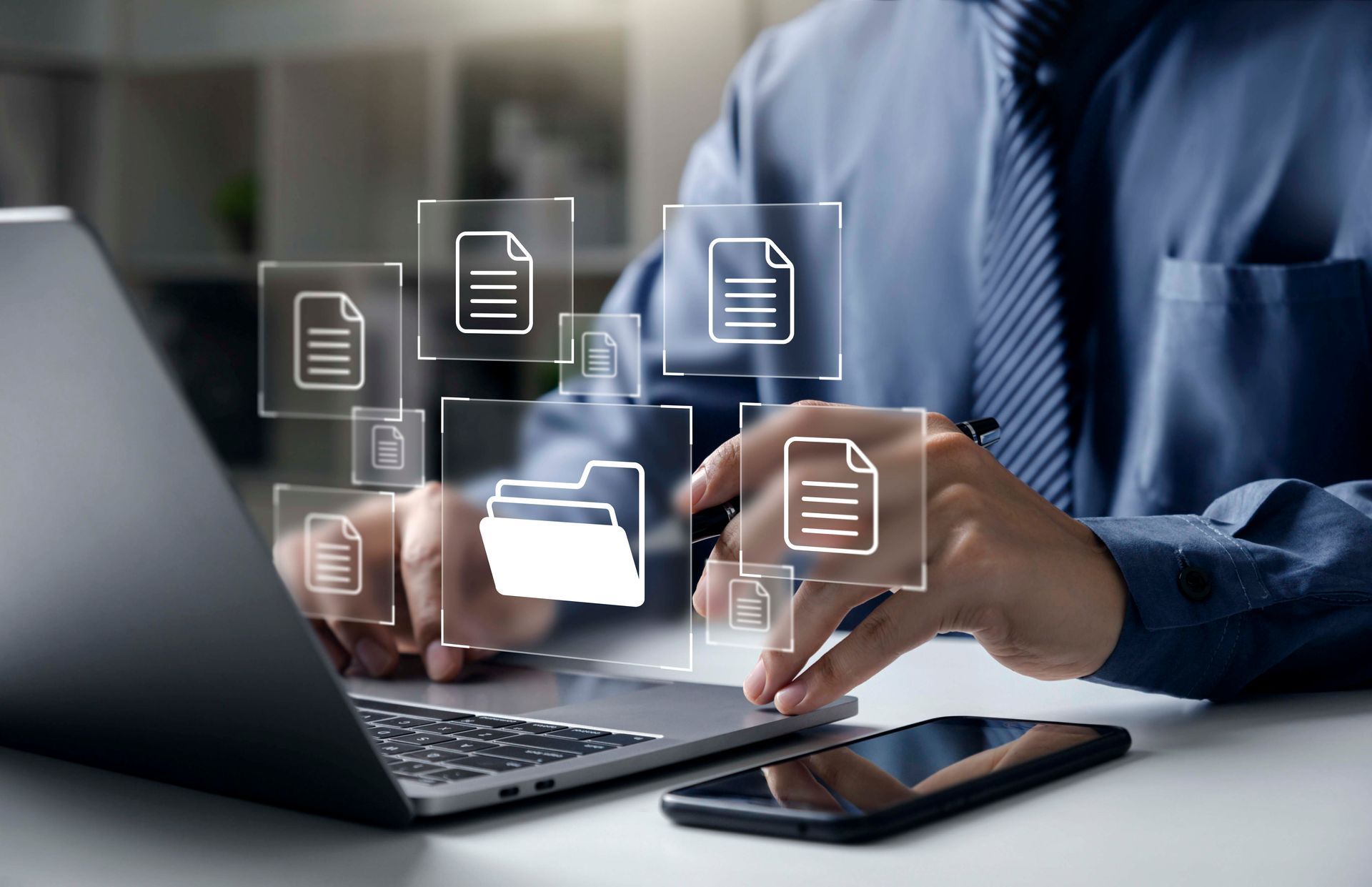 Person im blauen Hemd arbeitet am Laptop, über dem digitale Dateisymbole eingeblendet sind. Das Telefon liegt auf dem Schreibtisch.