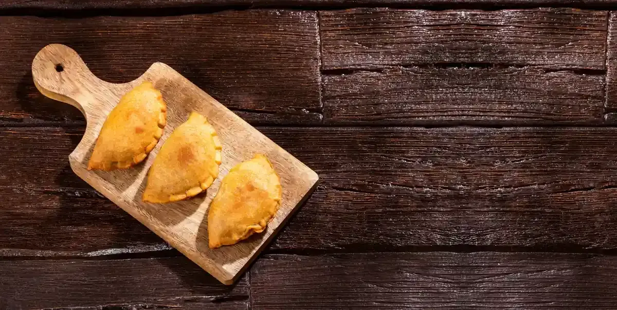 Tres empanadas de color marrón dorado sobre una tabla de cortar de madera, colocada sobre una superficie de madera oscura.