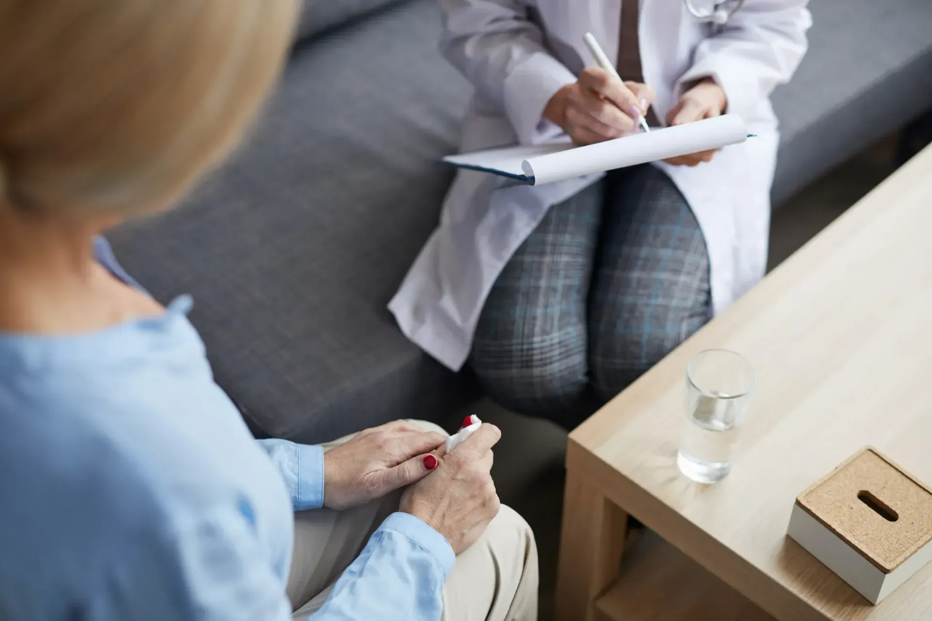 Una persona con camiseta azul consulta con un médico con bata blanca, ambos sentados. El médico toma notas.