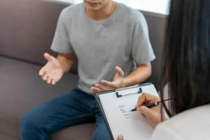 Hombre hablando con un terapeuta, sentado en un diván. El terapeuta toma notas en un portapapeles. Interior.
