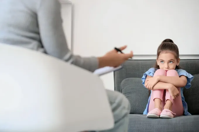 Un niño abraza sus rodillas en un sofá, mirando a una persona sentada con un portapapeles en una habitación luminosa.