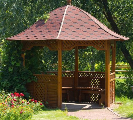En un jardín soleado, rodeado de vegetación y un pequeño rosal, se alza una glorieta de madera con techo de tejas rojas.