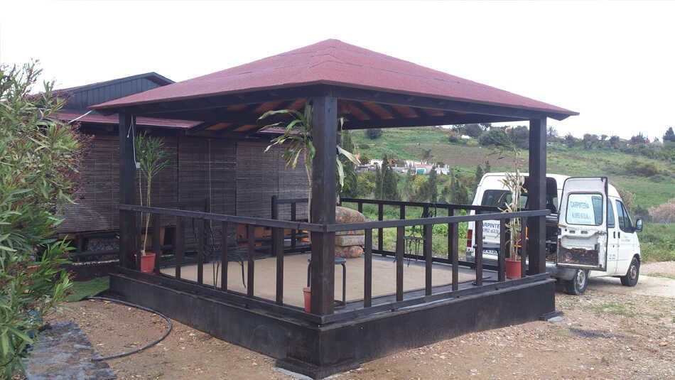 Un cenador de madera con estructura oscura y techo rojo se alza al aire libre junto a una furgoneta blanca en un terreno de tierra.