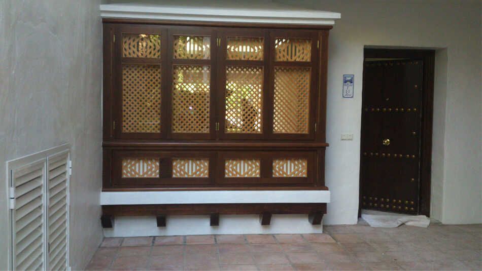 Un espacio exterior con un ventanal de celosía de madera oscura y una puerta a juego, que contrasta con las paredes blancas y los suelos de baldosas.