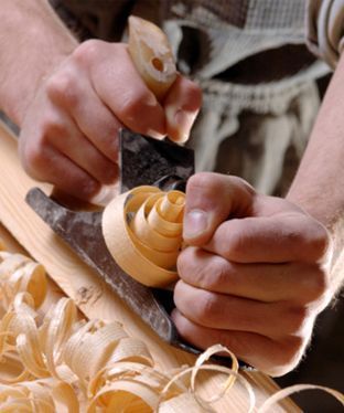 Una persona utiliza un cepillo de carpintero manual para desbastar una espiral de madera de una tabla, rodeada de virutas de madera.