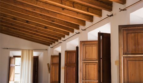 Una habitación interior con techos inclinados con vigas de madera, paredes blancas y contraventanas de madera abiertas sobre varias ventanas.