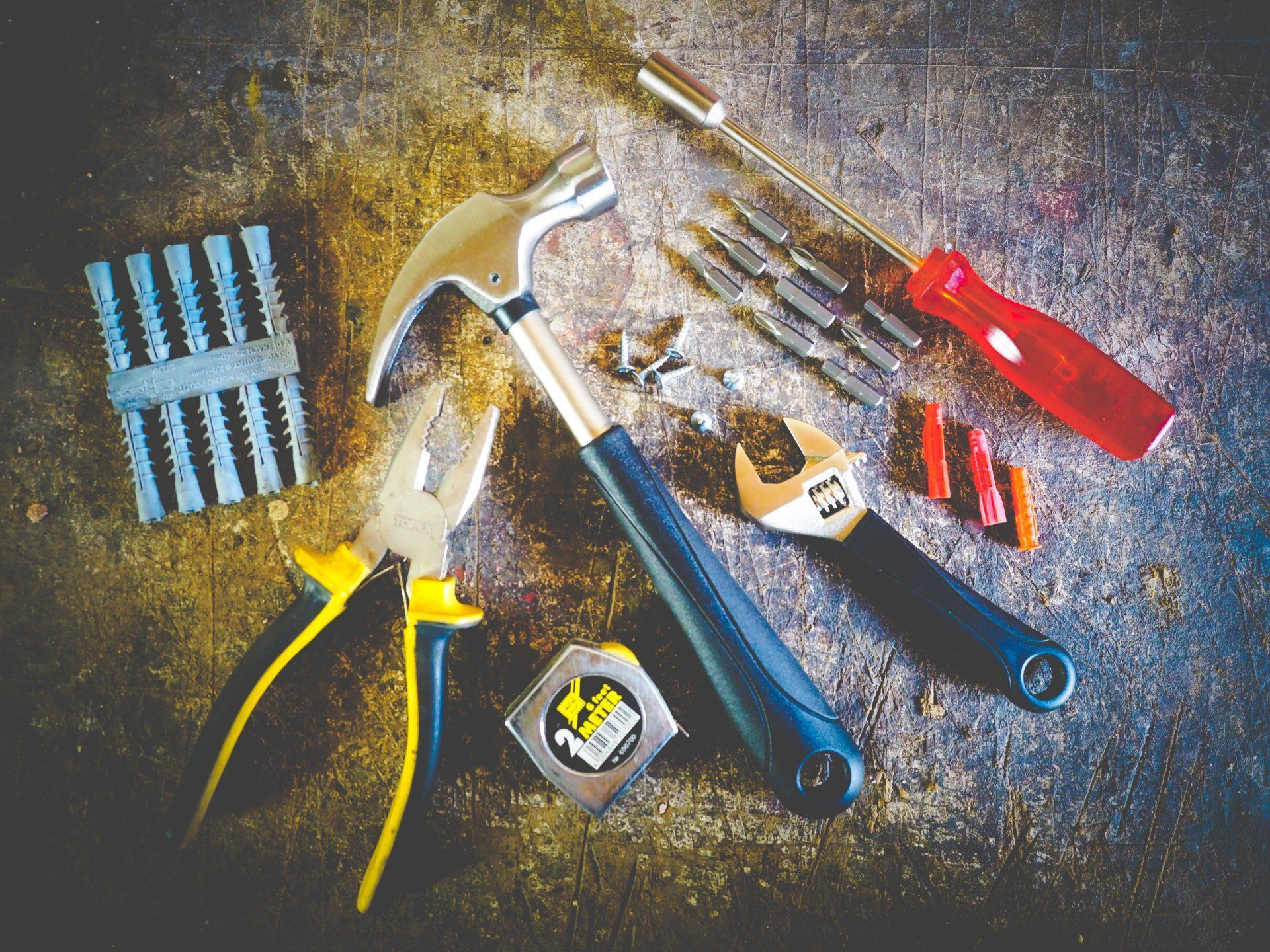 Verschiedene Handwerkzeuge auf einer abgenutzten Werkbank: Hammer, Schraubendreher, Zange, Schraubenschlüssel und Massband.