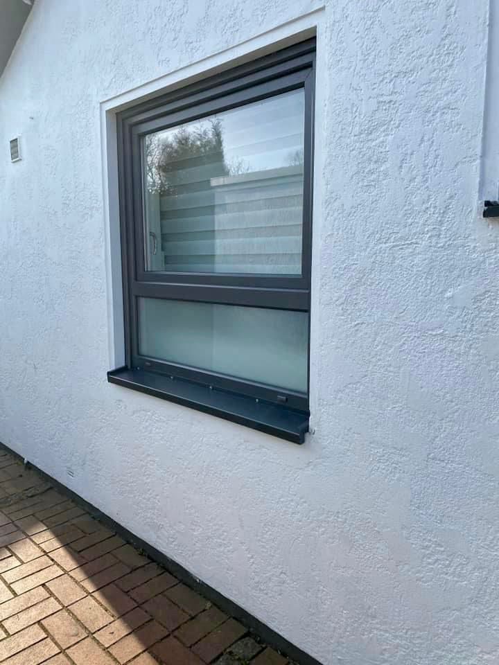 Fenster mit dunkelgrauem Rahmen an weißer Stuckwand, Außenansicht. Darunter eine Ziegelterrasse.