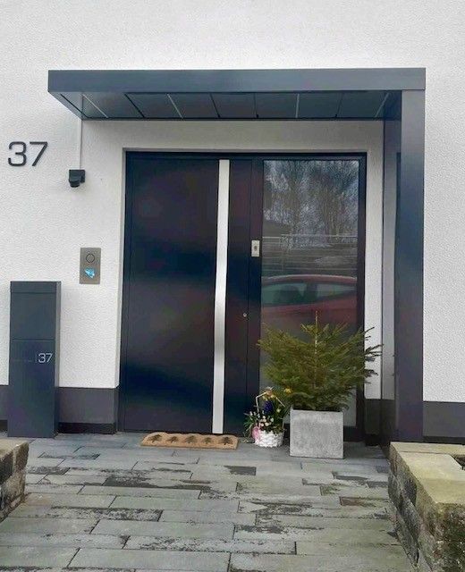 Moderner Hauseingang mit dunkler Tür, Glaspaneel und grauer Veranda. Hausnummer 37 ist sichtbar.