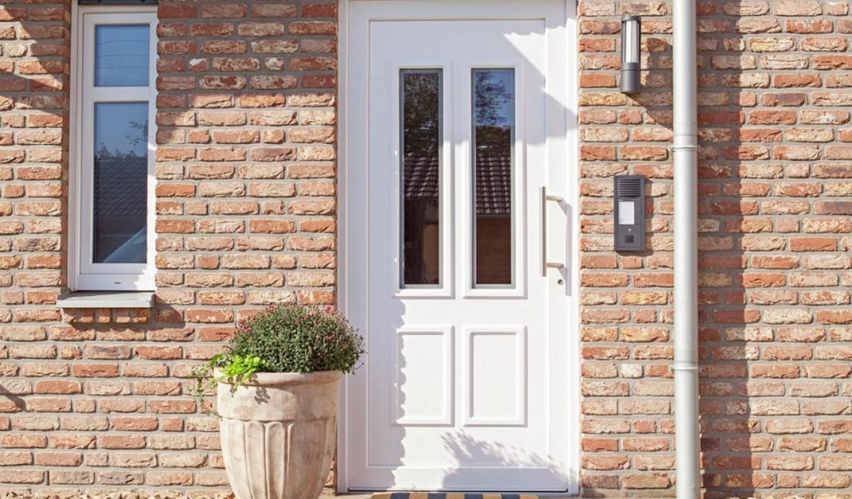 Weiße Haustür mit zwei Glasscheiben und zwei unteren Paneelen an einer Backsteinmauer, daneben eine Topfpflanze.