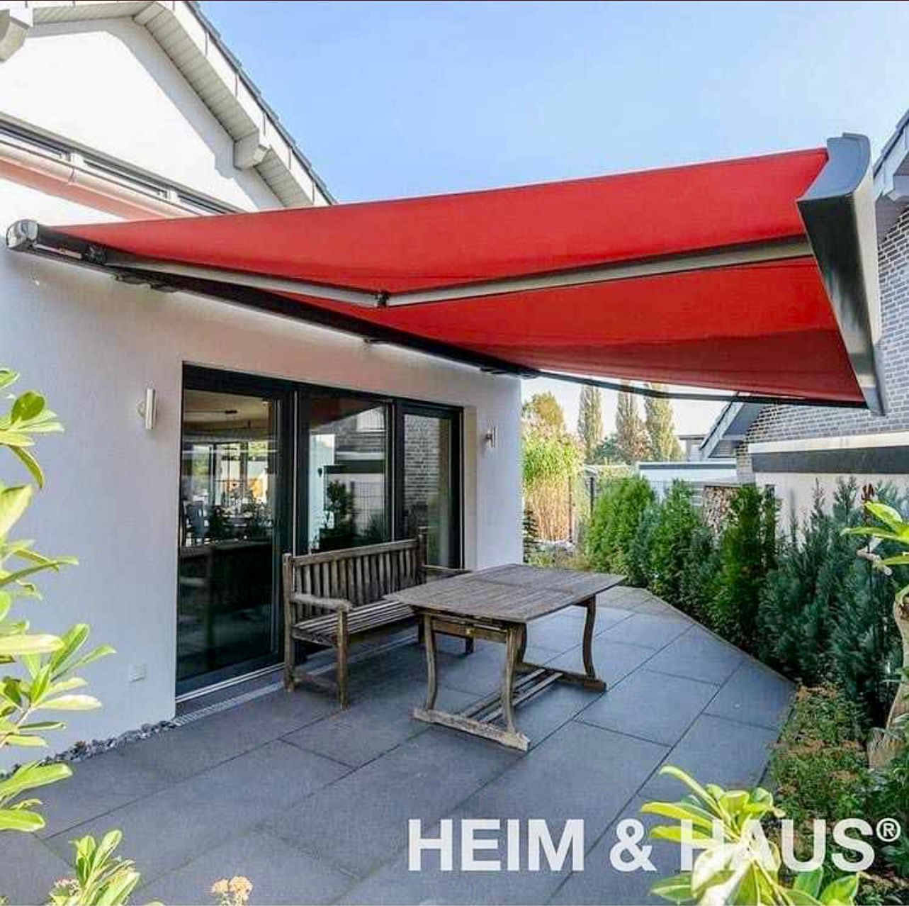 Eine rote Markise überspannt eine Terrasse mit Holztisch, Bank und Pflanzen.