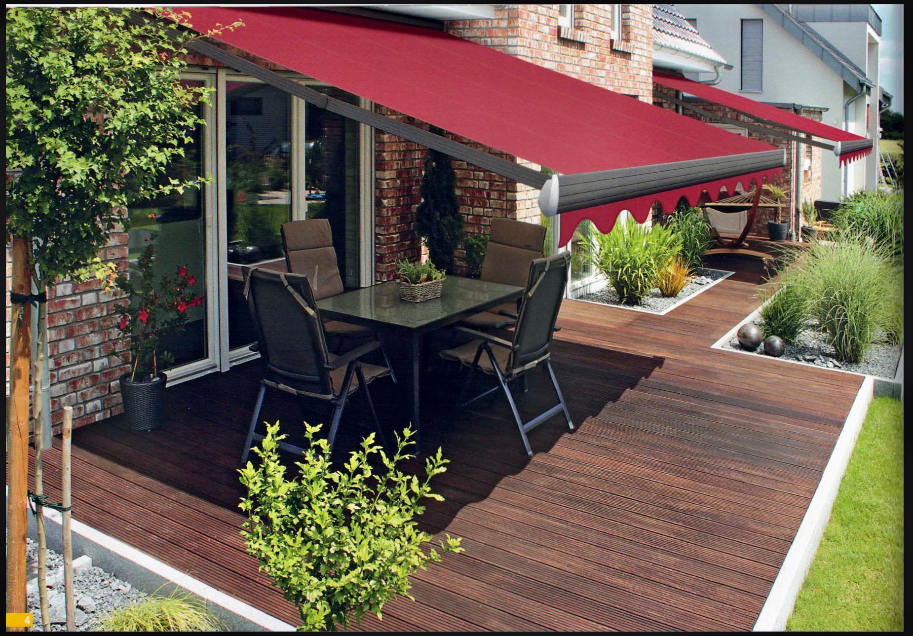 Eine rote Markise spendet Schatten auf einer Terrasse mit dunklen Möbeln. Backsteinhaus, Holzterrasse, Garten.