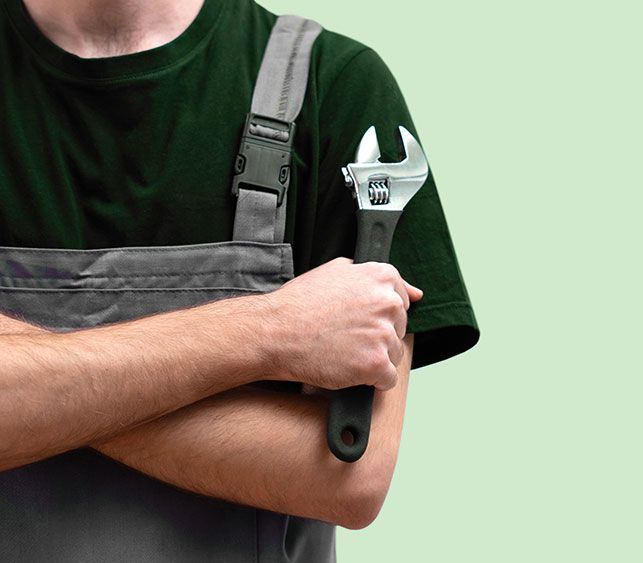 Mann in grünem Hemd und Latzhose, der einen Schraubenschlüssel hält, die Arme verschränkt.