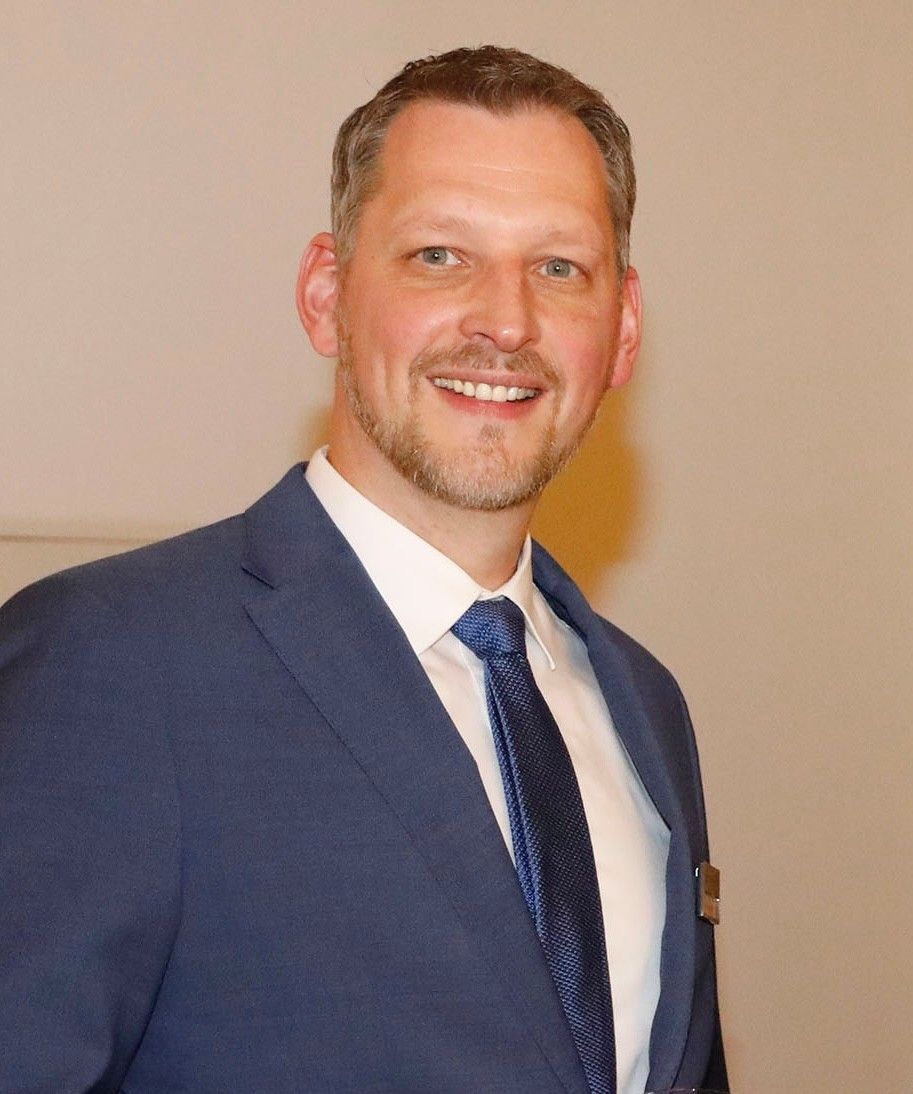 Mann im blauen Anzug, lächelnd. Er steht vor einer neutralfarbenen Wand und trägt eine gemusterte blaue Krawatte.