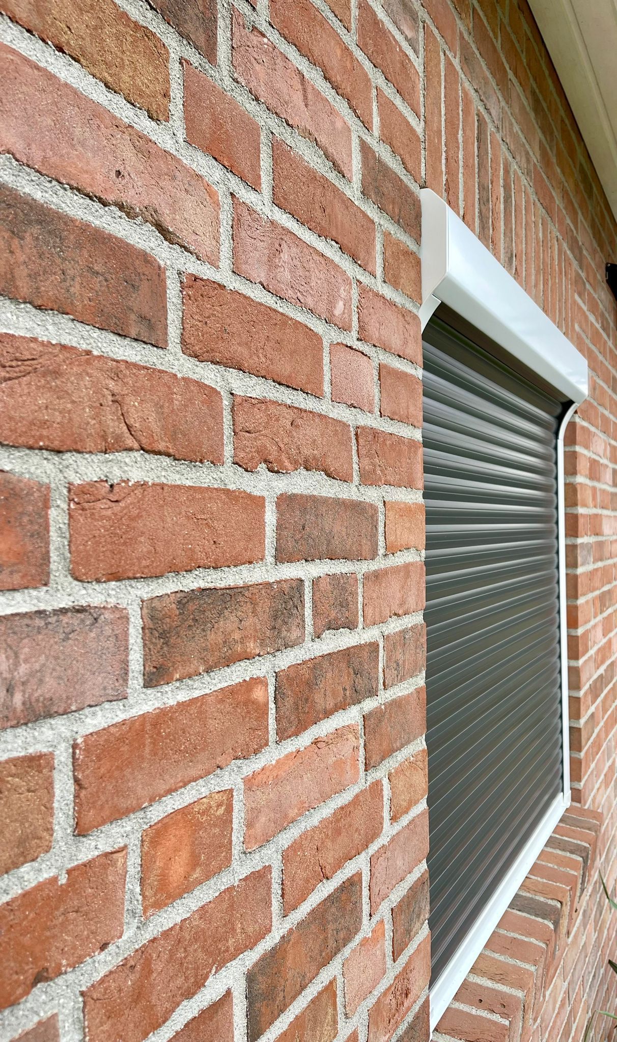 Außenansicht einer Backsteinmauer mit einem grauen, geschlossenen Rollladen über einem Fenster.