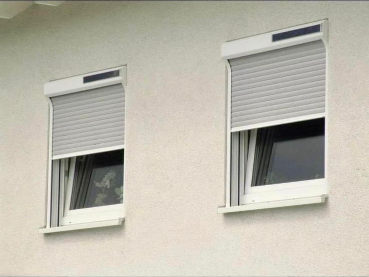 Zwei Fenster mit grauen Rollläden an einer hellen Gebäudefassade.