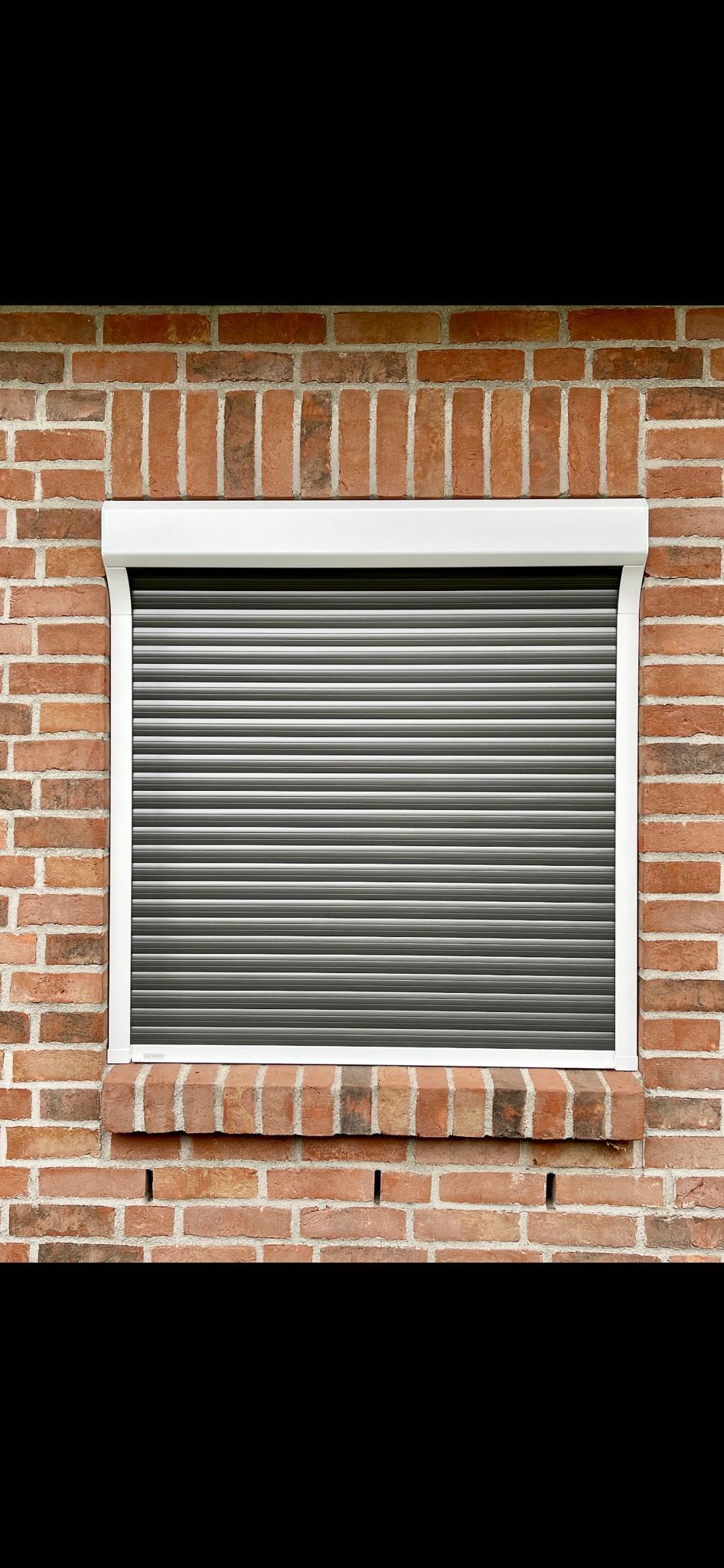 Fenster mit geschlossenen Sicherheitsrollläden aus Metall, eingelassen in eine Ziegelwand. Weißer Rahmen und Sturz.