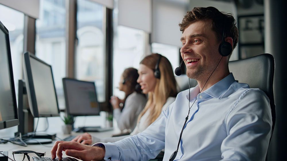 Ein Mann mit Headset sitzt lächelnd am Computer und arbeitet mit anderen in einem Büro.