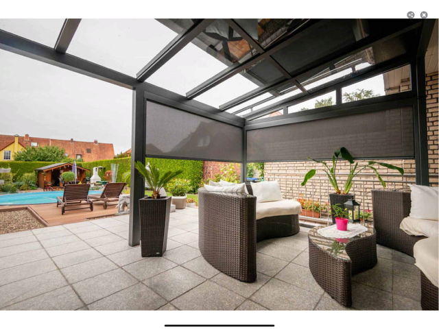 Terrasse mit dunkelgrauer Überdachung, Gartenmöbeln und Sonnenschutz. Pool und Grünanlagen im Hintergrund.