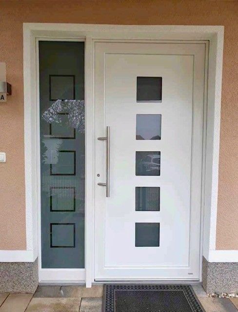 Weiße Haustür mit quadratischen Glasscheiben und einer Seitenwand, silbernem Türgriff und schwarzer Fußmatte.