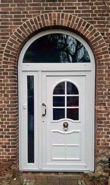 Weiße, bogenförmige Eingangstür mit Seitenwand, eingelassen in eine Backsteinfassade.