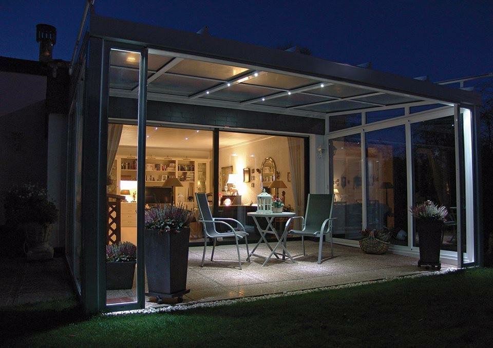 Beleuchteter Terrassenbereich bei Nacht, mit Tisch, Stühlen, Pflanzen und Blick in das beleuchtete Innere eines Hauses.