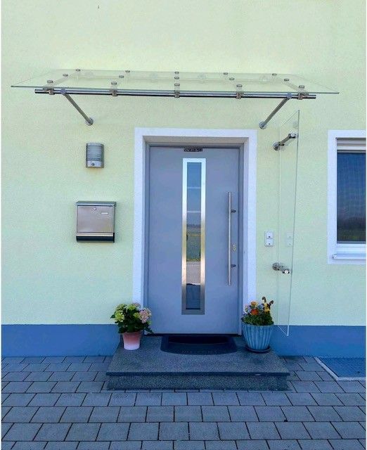 Hellgraue Eingangstür mit Glasvordach und Seitenwand; hellgelbes Haus.