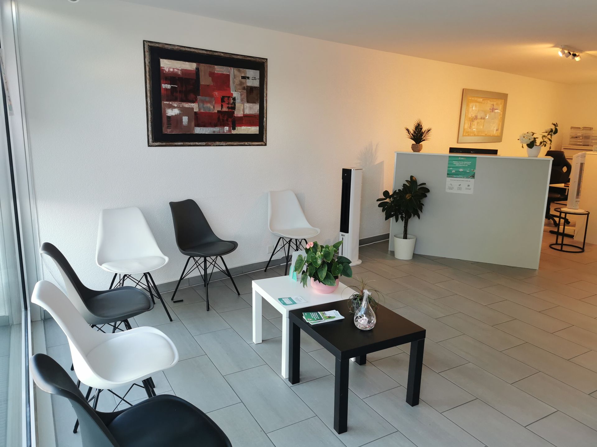 Salle d'attente lumineuse avec chaises blanches et noires, petites tables, plantes et comptoir d'accueil.